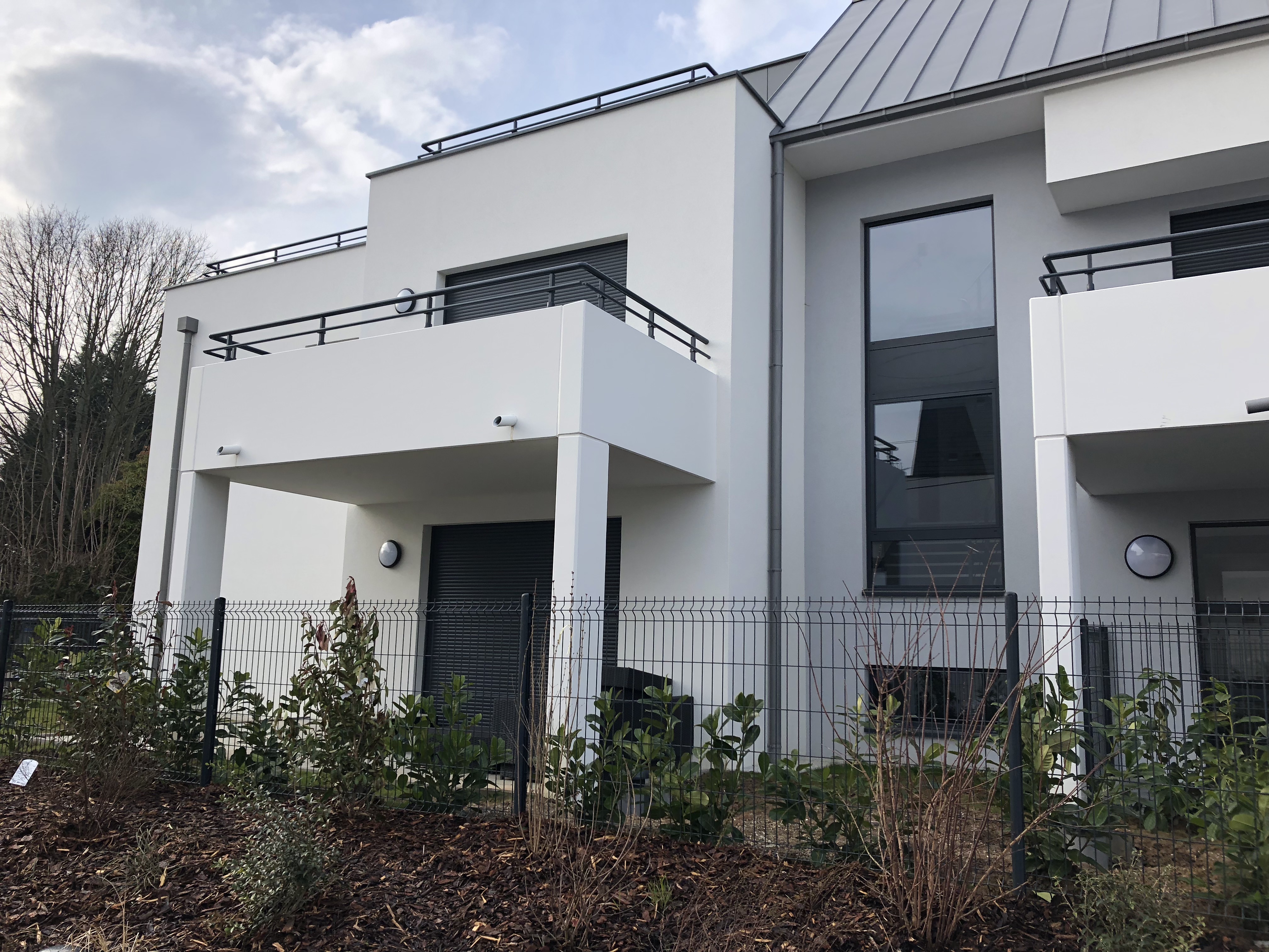 Vue d'une maison moderne avec balcons, murs blancs et fenêtre large, entourée d'un jardin et d'une clôture.