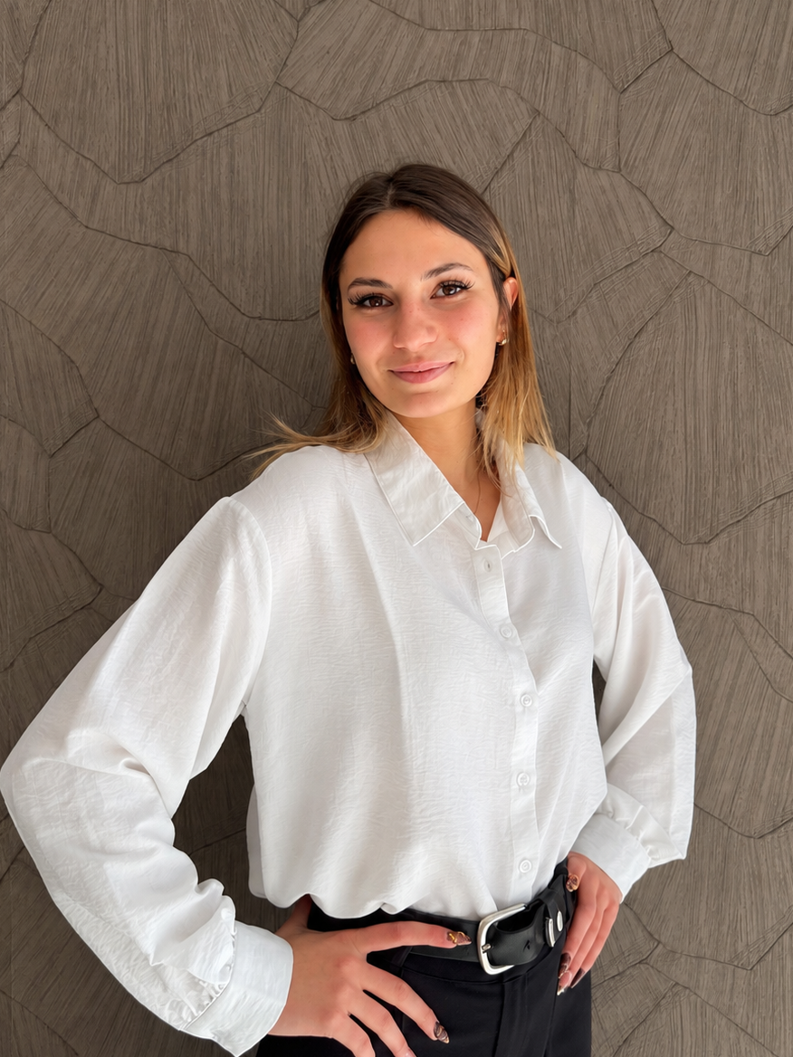 Une jeune femme souriante posant devant un mur texturé, portant une chemise blanche et un pantalon noir.
